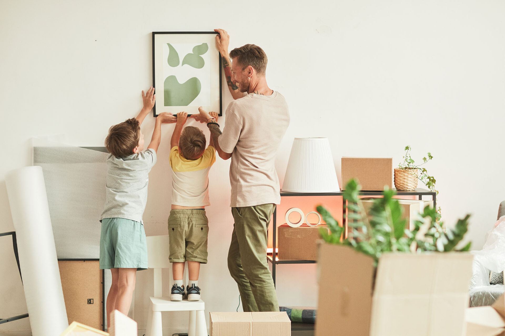 Full length portrait of happy father with two sons hanging pictures on wall while moving in to new home, copy space